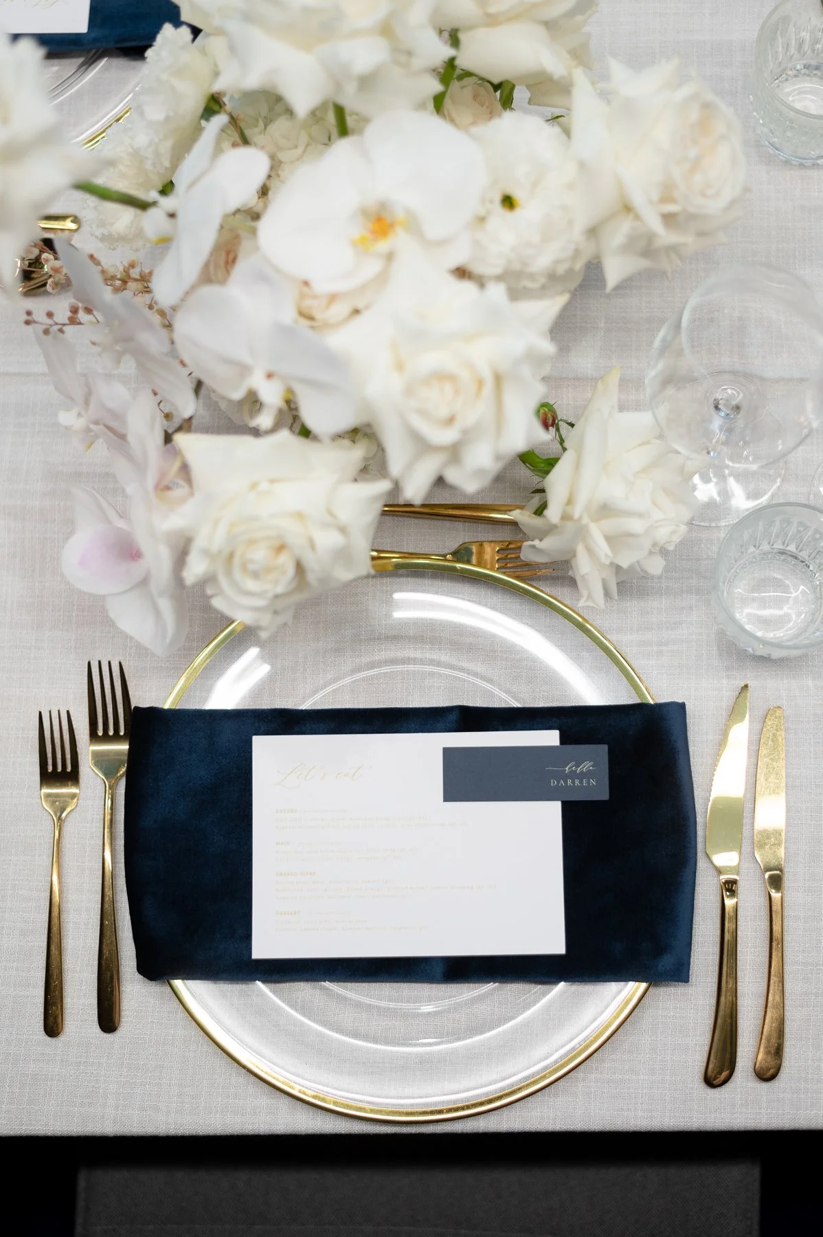 Elegant place setting with a clear glass charger plate, gold flatware, navy napkin, wedding menu, and a floral centerpiece of white flowers on a table with a light-colored tablecloth.