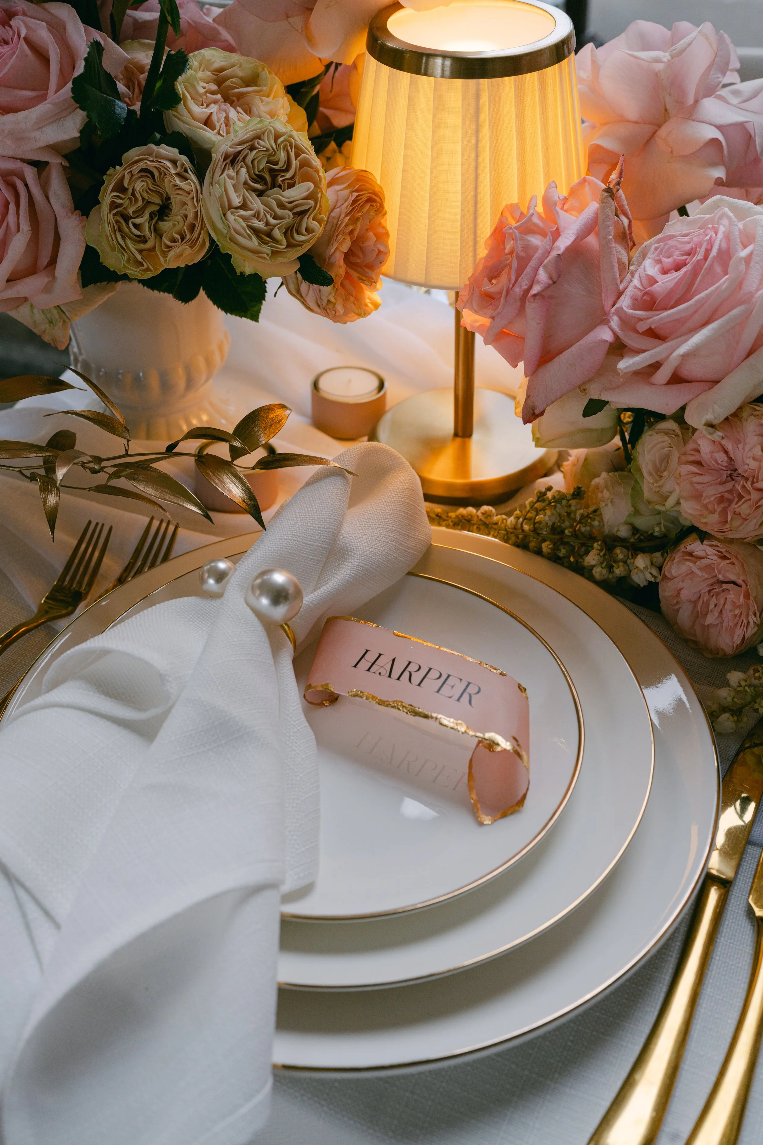 Elegant table setting with a white plate, gold utensils, a pink name card with 'HARPER,' a white napkin with pearl napkin ring, surrounded by pink and cream roses, a gold lamp, and a small candle.