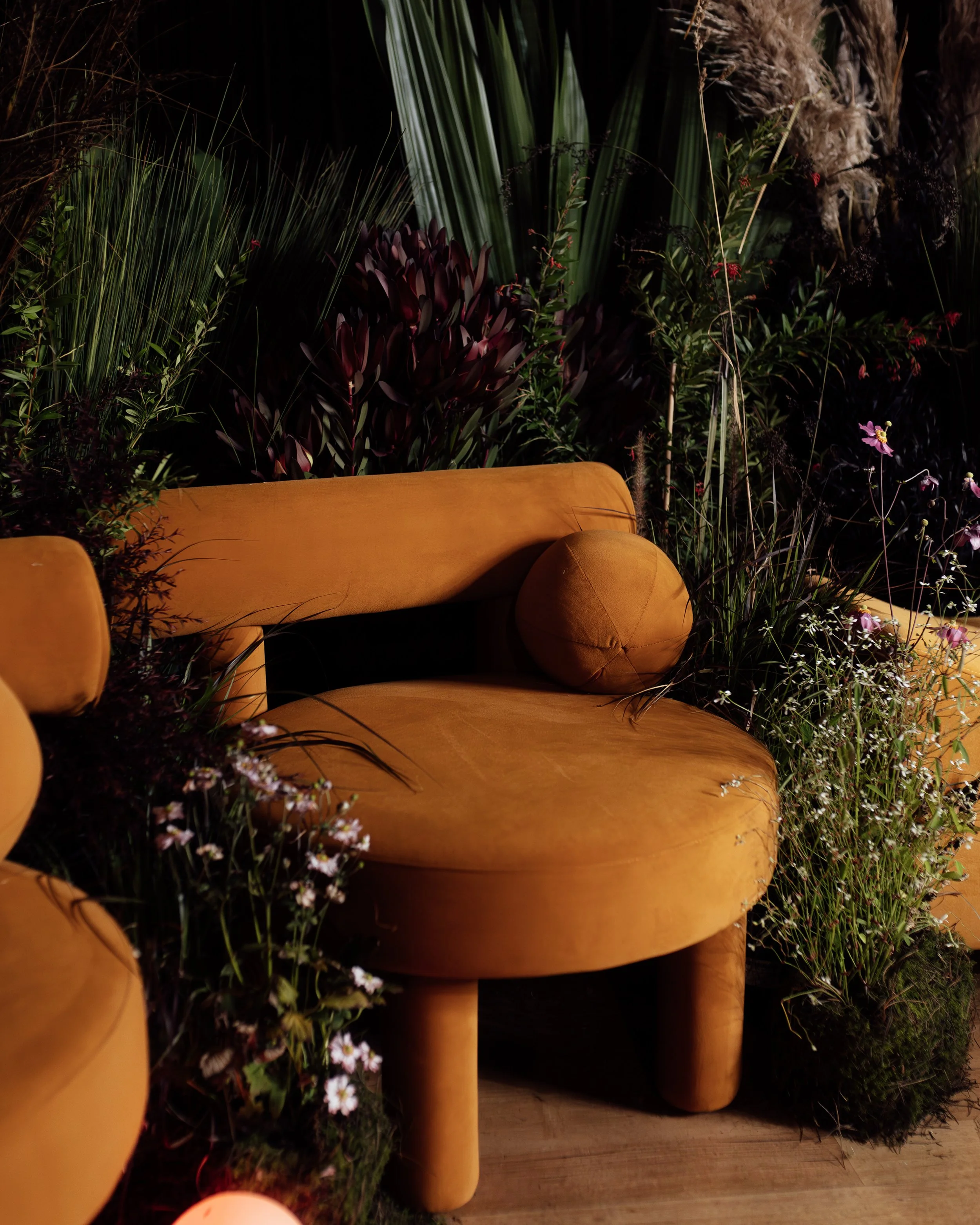 A corporate stage panel discussion setting for a high end elevated experience using premium furniture. A mustard-colored velvet sofa decorated with a round pillow, surrounded by lush green plants and flowers in a dimly lit setting.