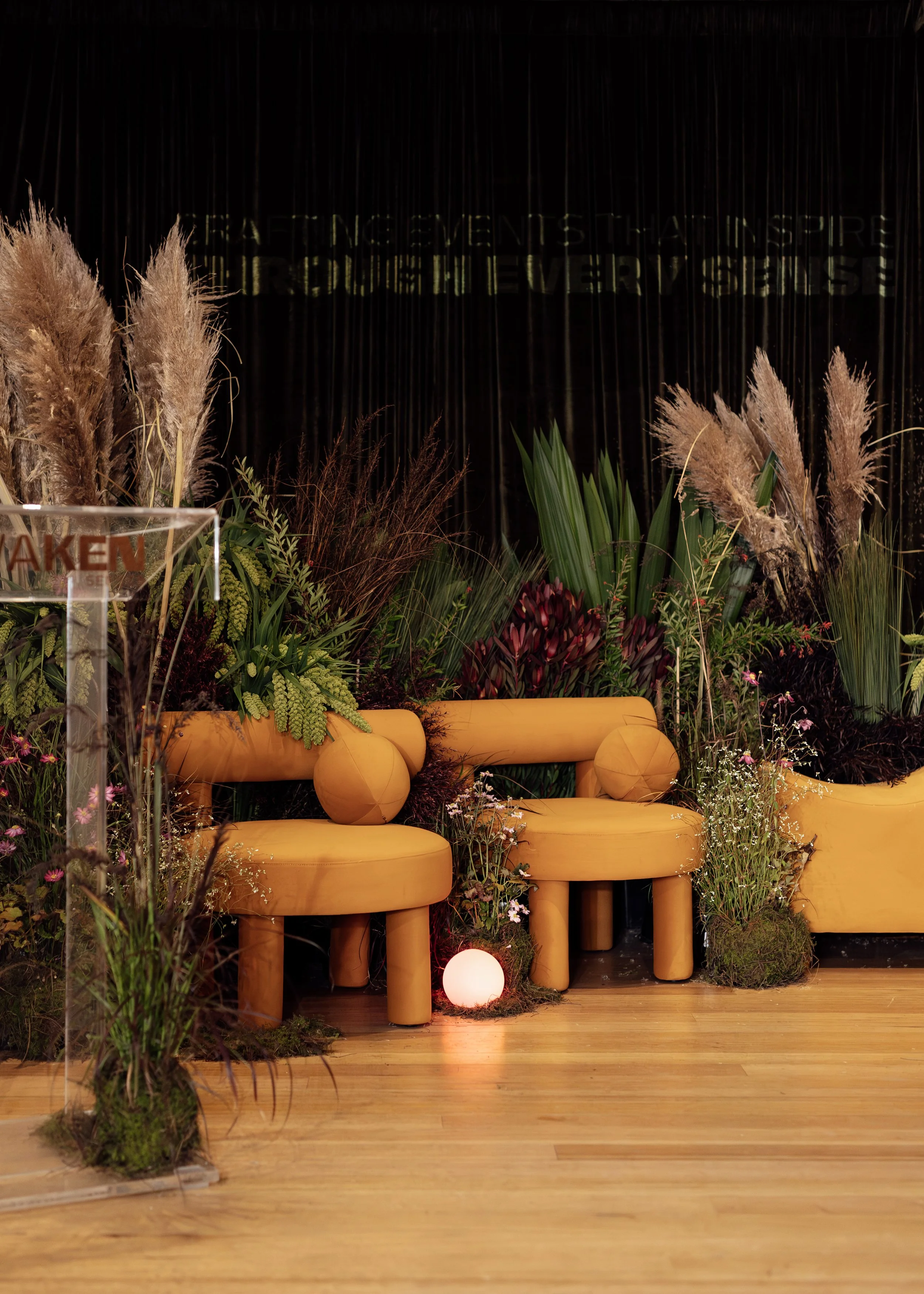 Decorative indoor event setting with yellow padded chairs surrounded by lush plants and flowers, with a black backdrop and glowing round light on the floor.