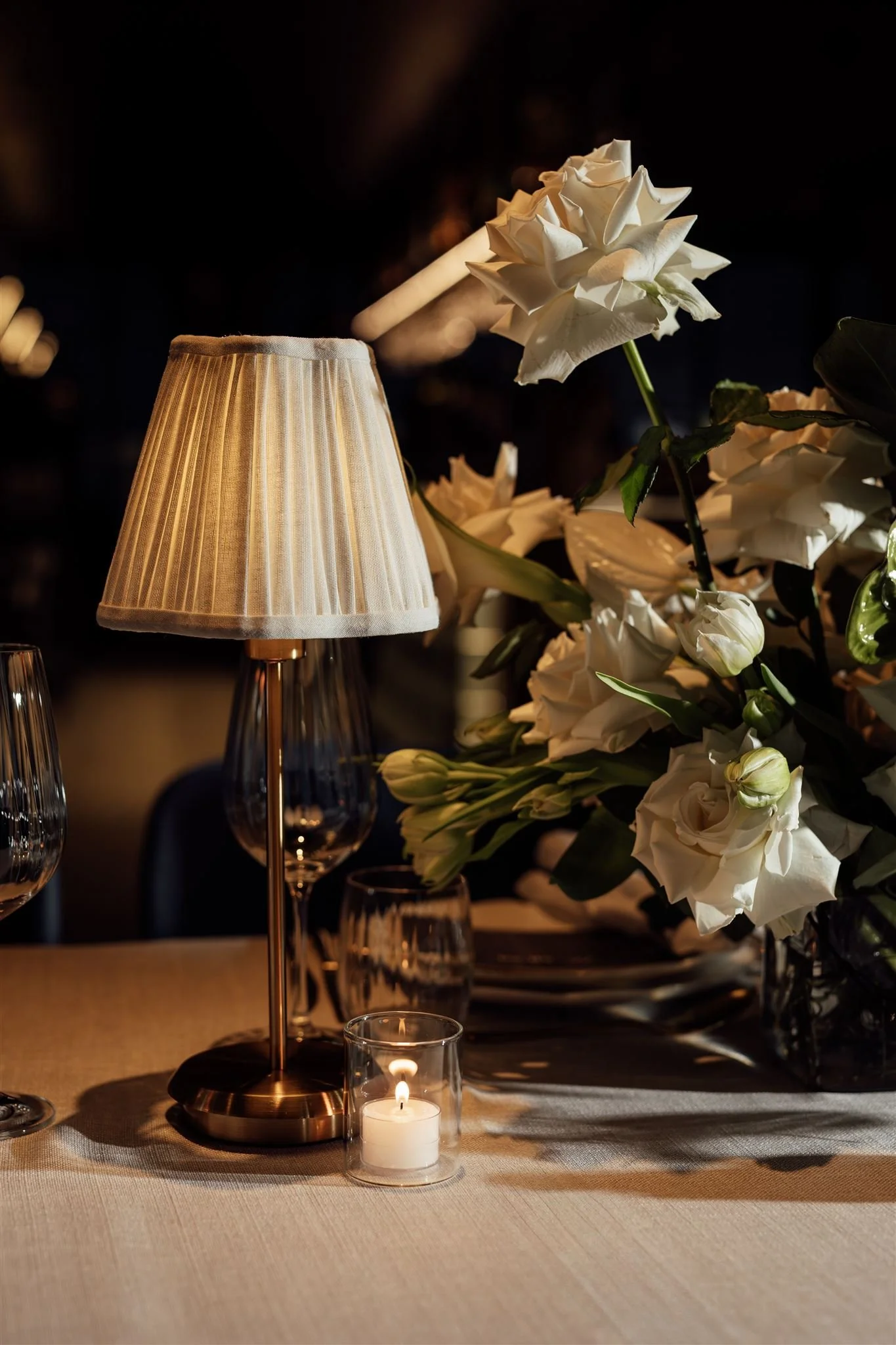 A table setting with a small table lamp, a lit candle in a glass holder, and a flower arrangement of white roses and lilies.