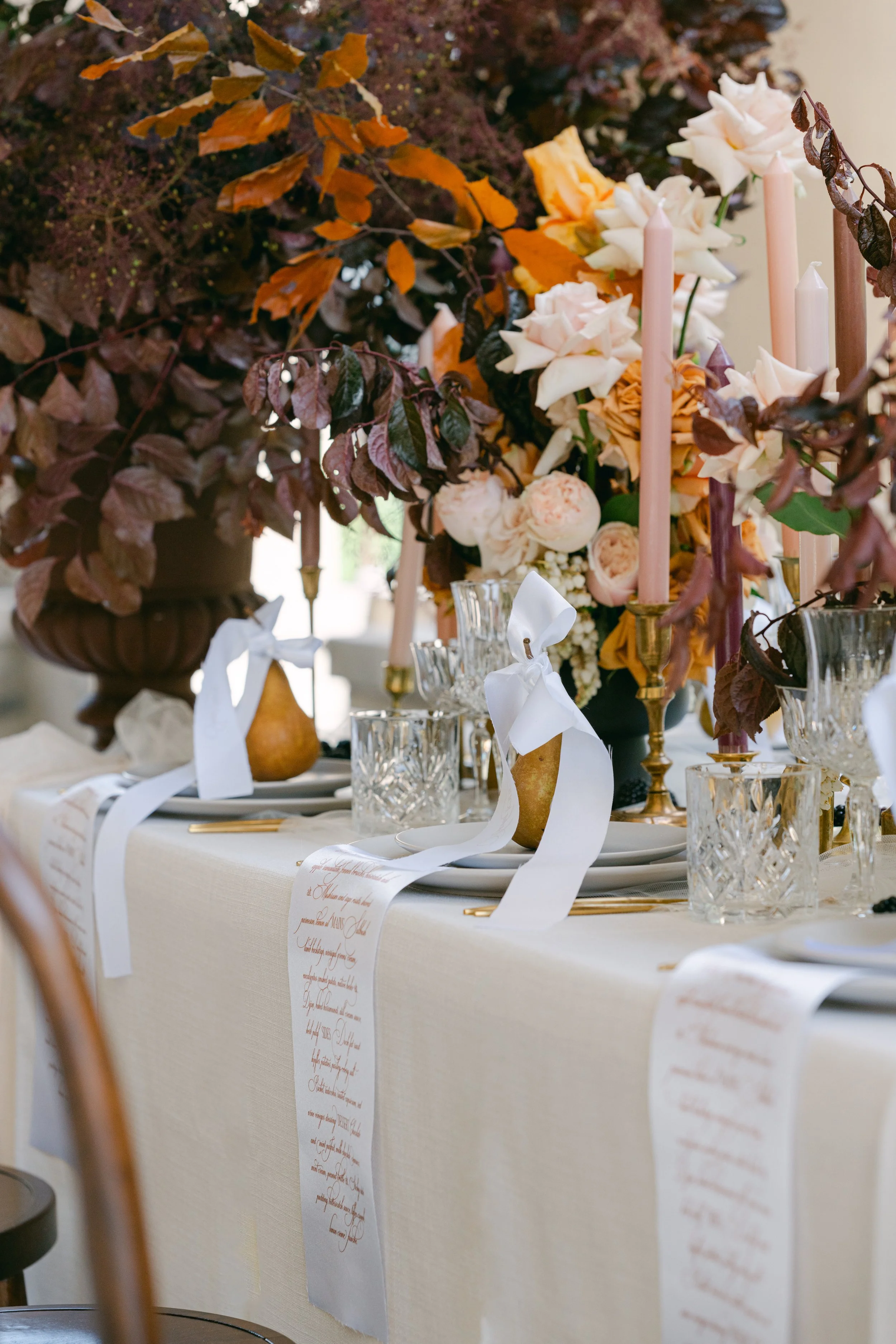 Elegant table setting with tall pink candles, floral arrangements with white and peach flowers, crystal glassware, white plates, and white ribbons tied to pears, with a scroll-like menu hanging over the table edge.