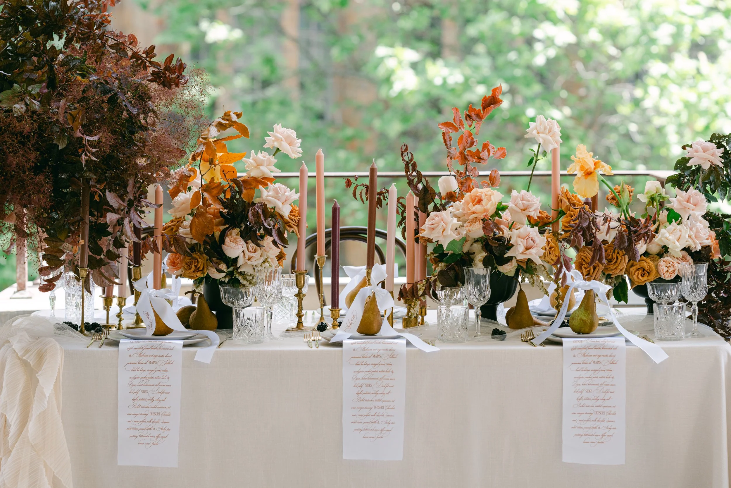A beautifully decorated dining table with peach, cream, and ochre flower arrangements, tall taper candles, glassware, and elegant place settings, set outdoors with green foliage in the background.