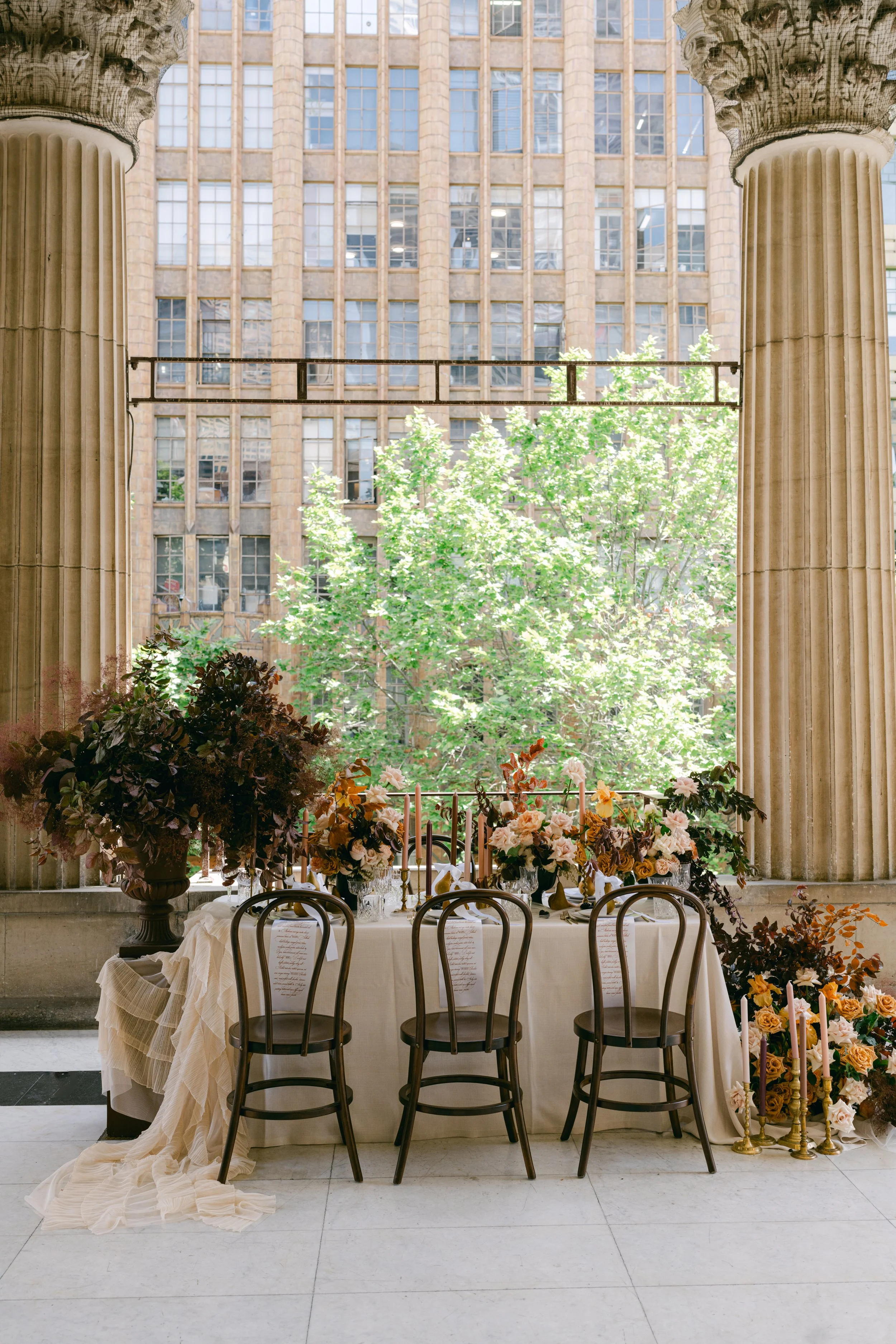 Elegant table setup with floral arrangements, candles, and chairs in front of large windows and stone columns