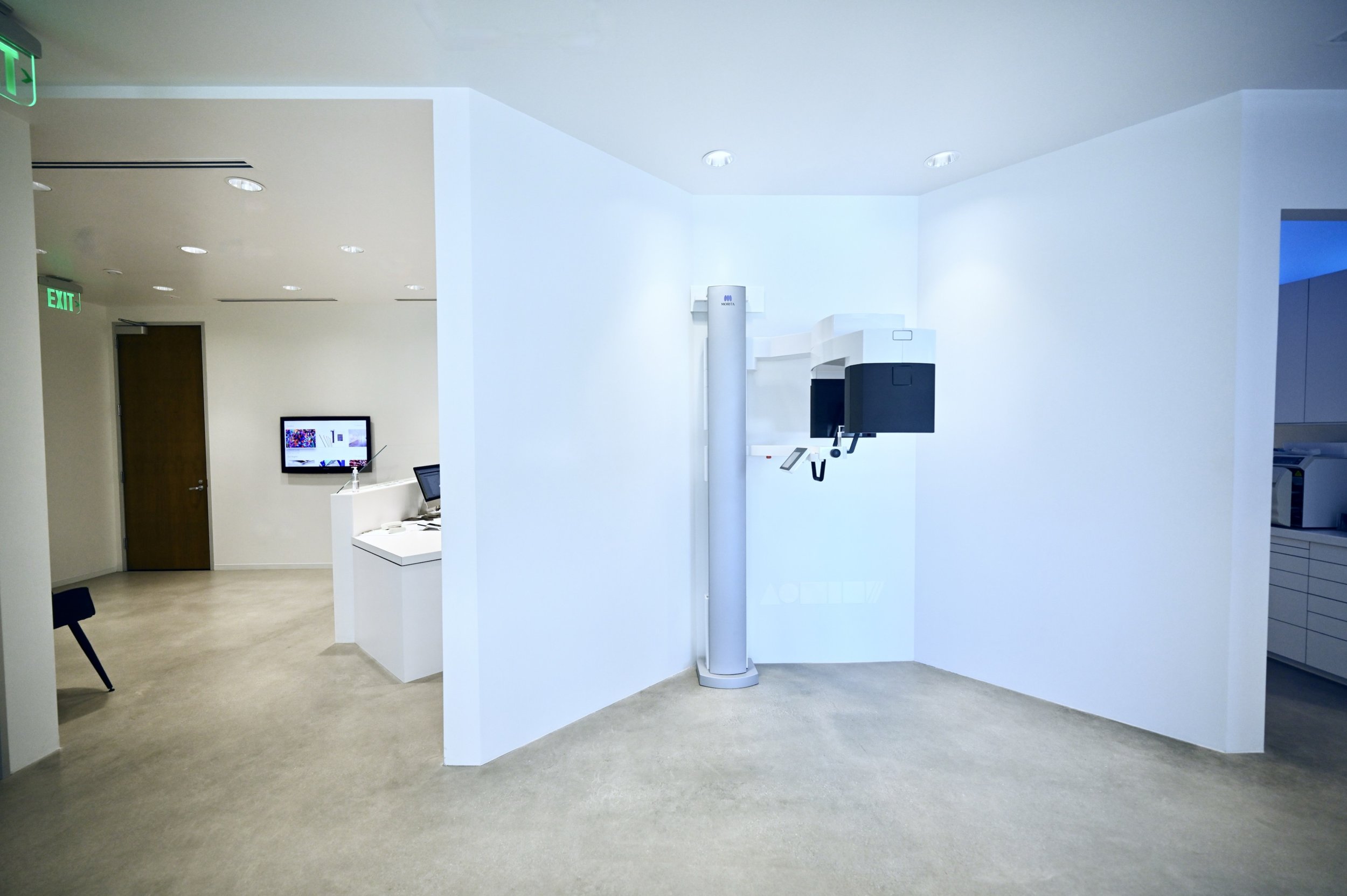 Medical or dental office interior with white walls, beige flooring, and modern equipment, including a tall white and black machine, monitors, and chairs.