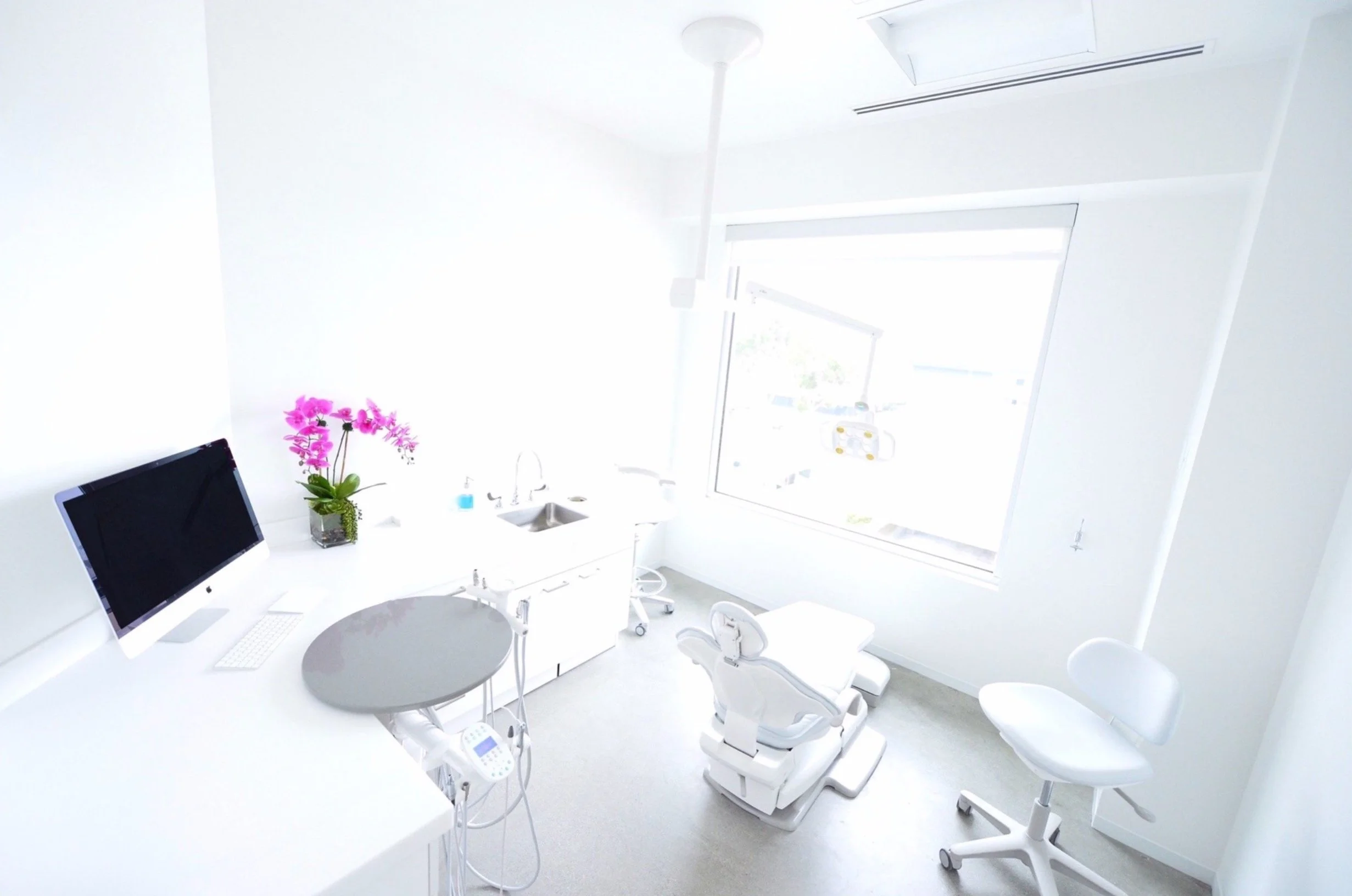 A modern dental clinic with white walls, dental chairs, a computer on the counter, a flower arrangement, and a window letting in natural light.