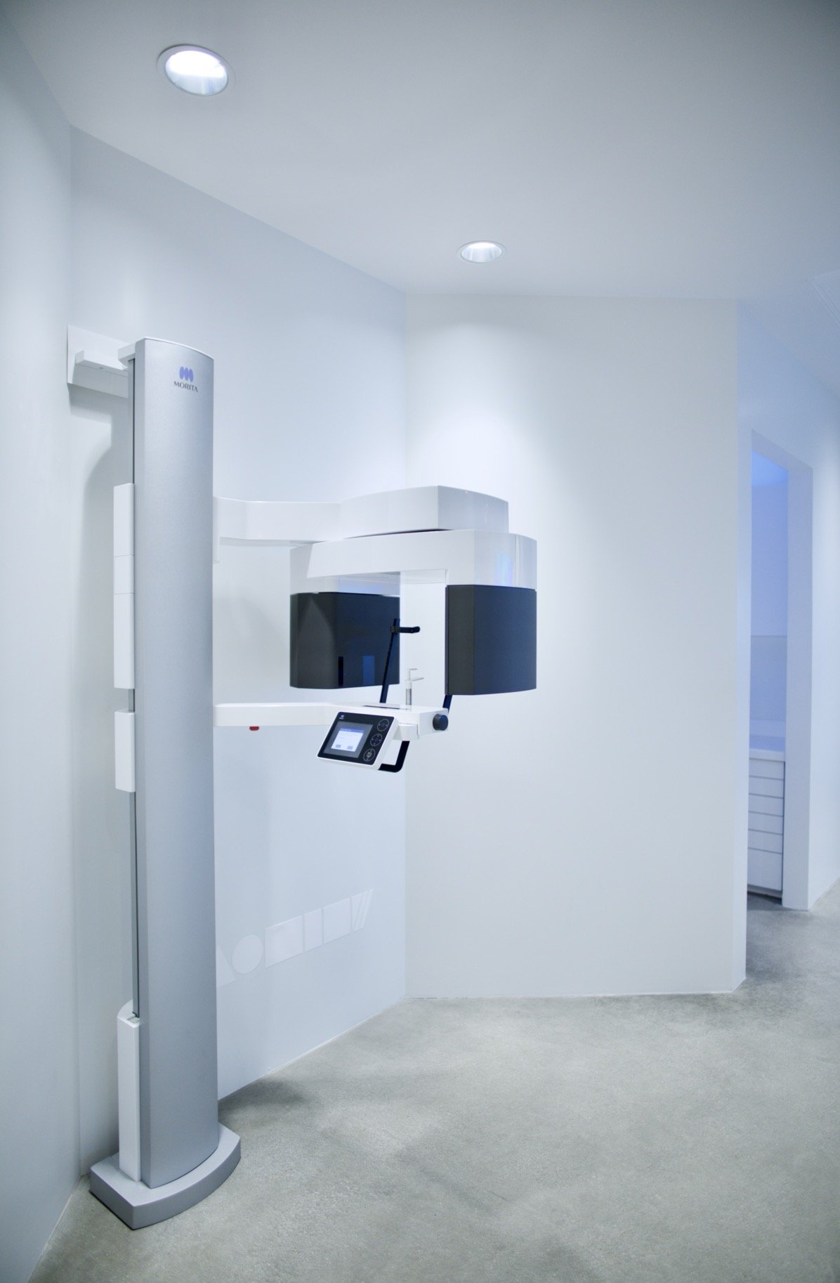 Medical imaging room with a panoramic X-ray machine, white walls, gray carpeted floor, and ceiling lights.