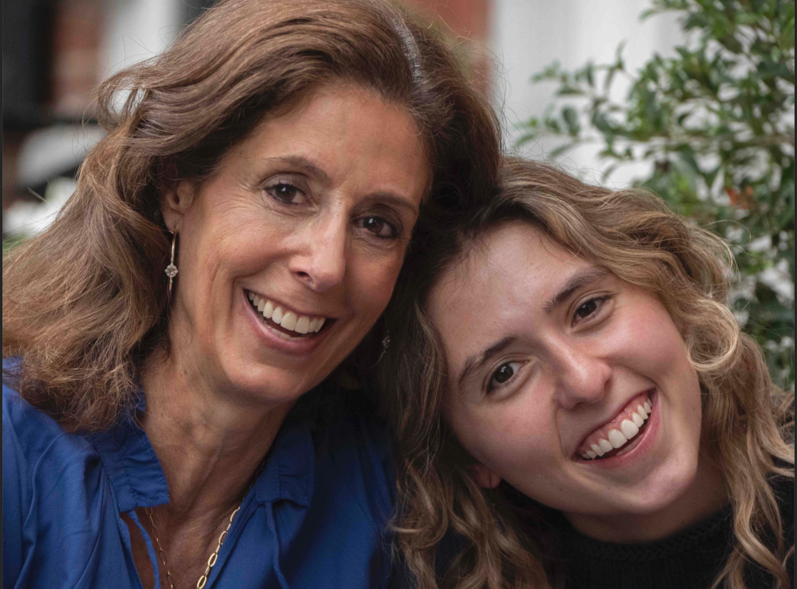 Two women smiling and leaning close together, with a background of greenery.