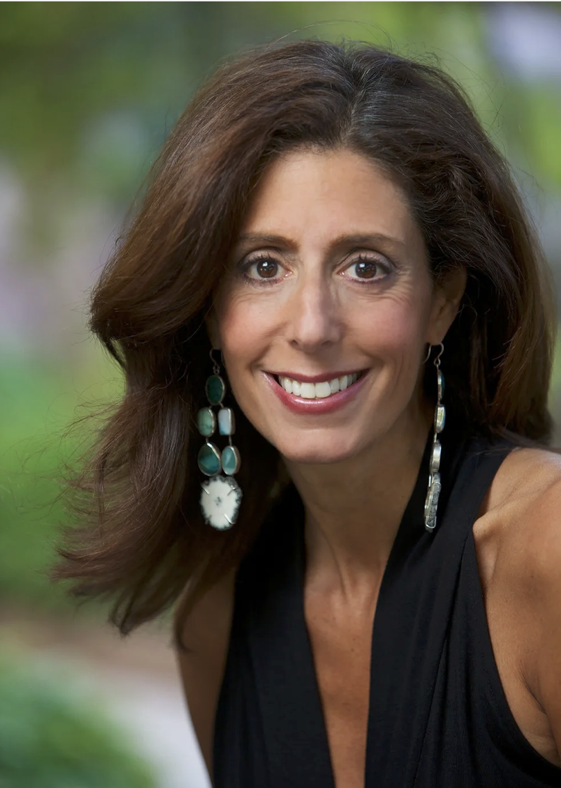 A woman with brown hair and brown eyes smiling outdoors, wearing a black top and large earrings with green and white stones.