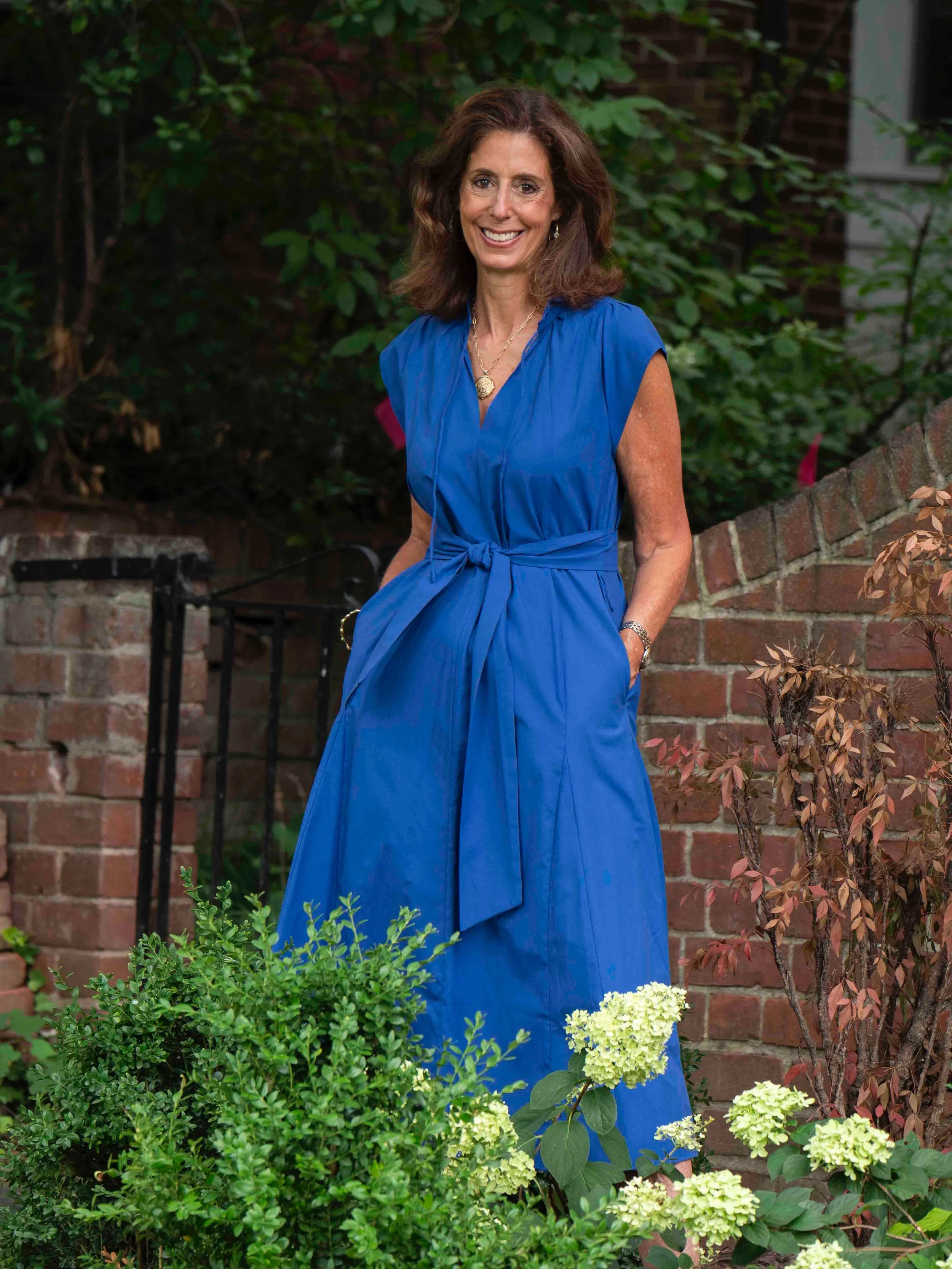 A woman with shoulder-length brown hair, wearing a bright blue dress and gold jewelry, smiles while standing outdoors in front of a brick wall and lush greenery and flowering plants.