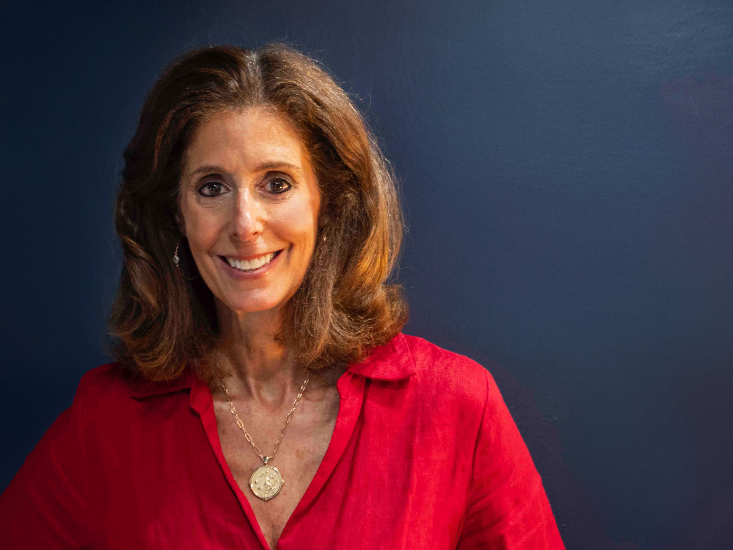 A smiling woman with brown hair wearing a red blazer, earrings, and a pendant necklace, against a dark blue background.