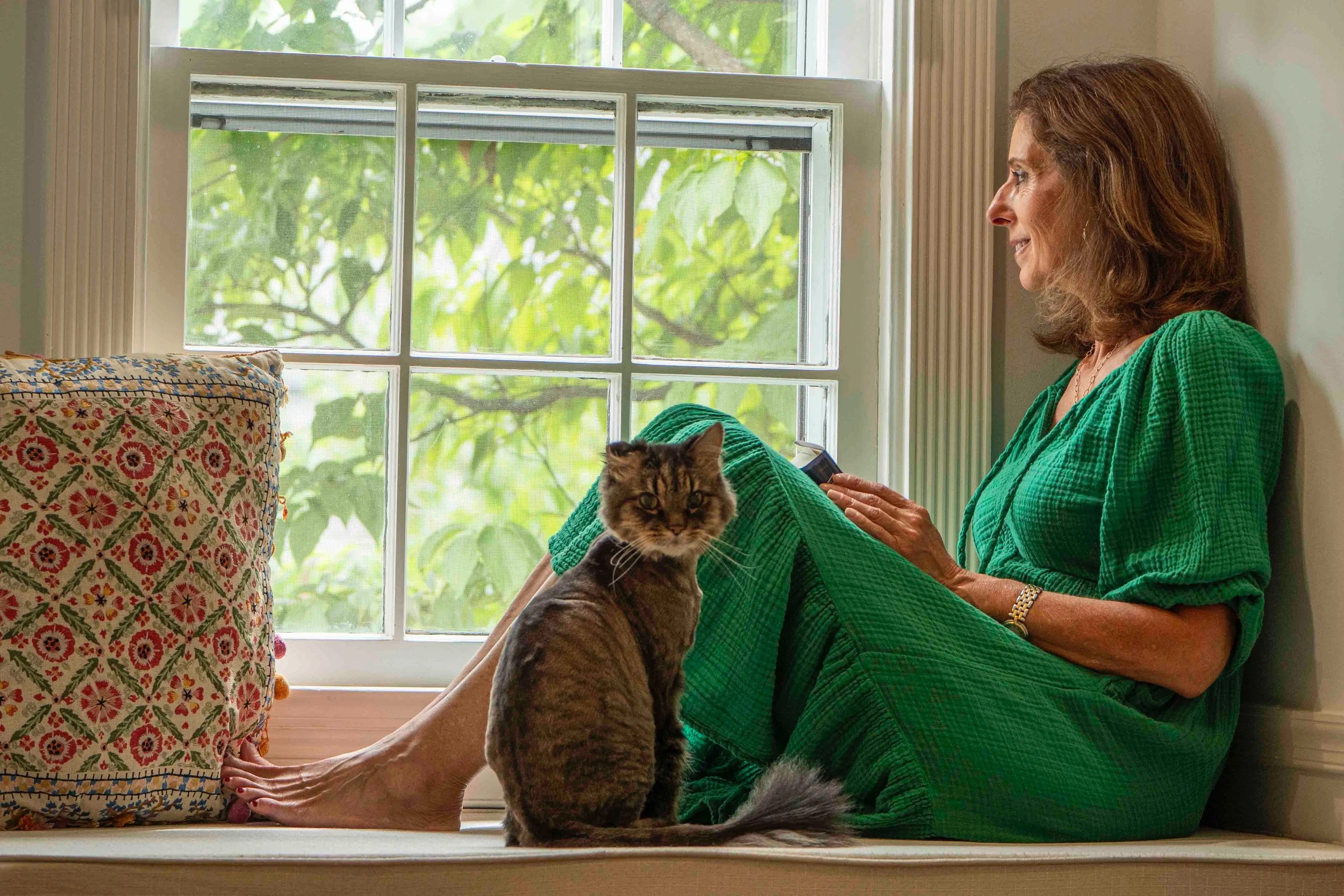 A woman in a green dress sitting on a windowsill with a cat on her lap. The woman has shoulder-length brown hair and is smiling while looking outside. There is a blanket with a floral pattern beside her and greenery visible outside the window.