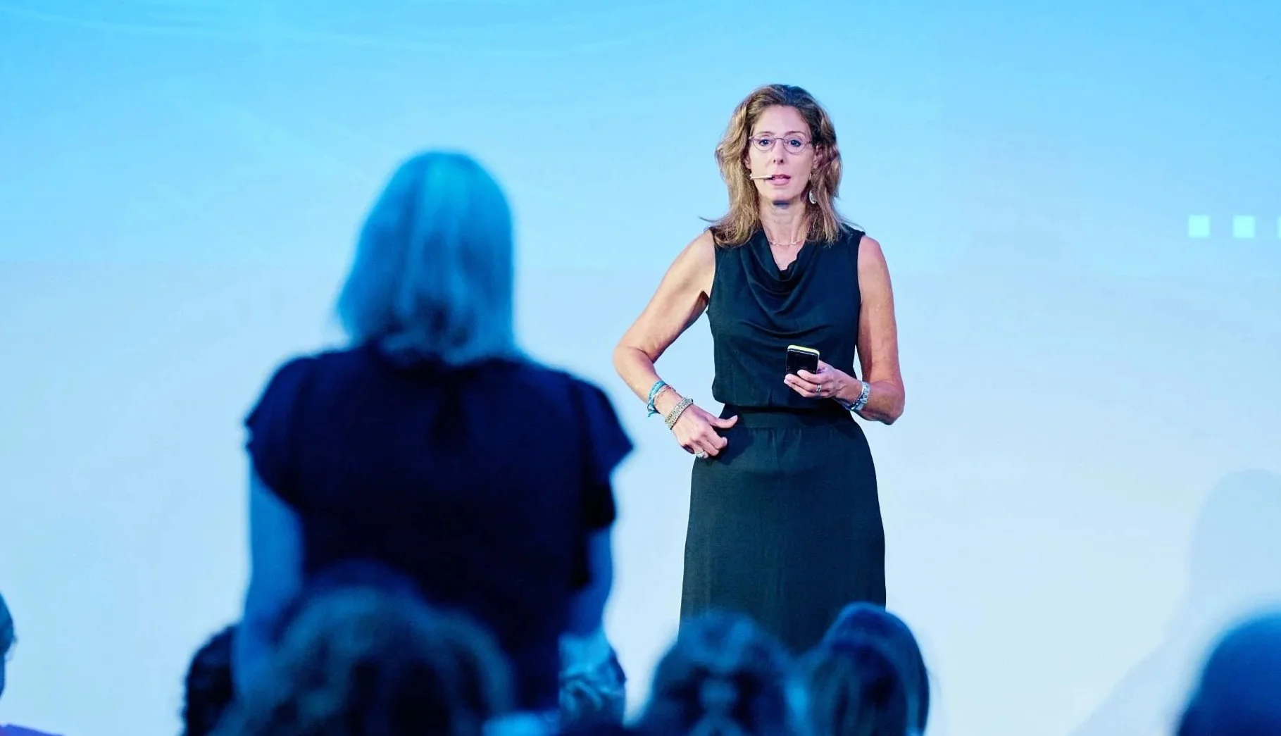 A woman in a black sleeveless dress speaking at a conference or event, holding a phone, with an audience in front of her.