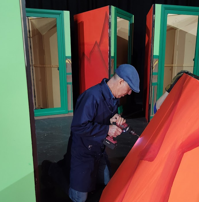 Man in blue coat and cap using a drill on a large red wooden prop with painted lines, surrounded by colorful stage set pieces resembling rooms or partitions.