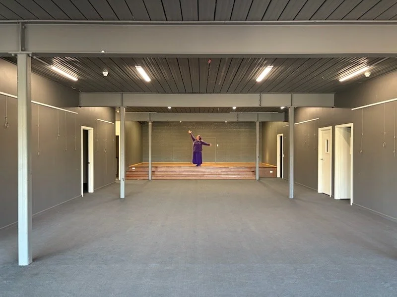 A woman in purple attire stands on a small stage inside a mostly empty, spacious room with gray walls and ceiling, and a gray carpeted floor, possibly practicing or performing a dance or yoga pose.
