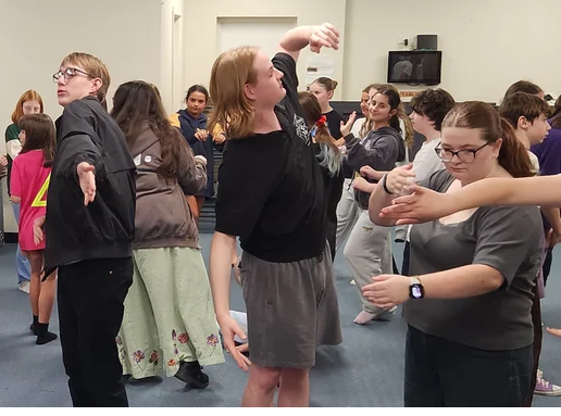 Group of young people dancing and interacting in a room, some with their eyes closed or looking around, with a speaker on the wall.