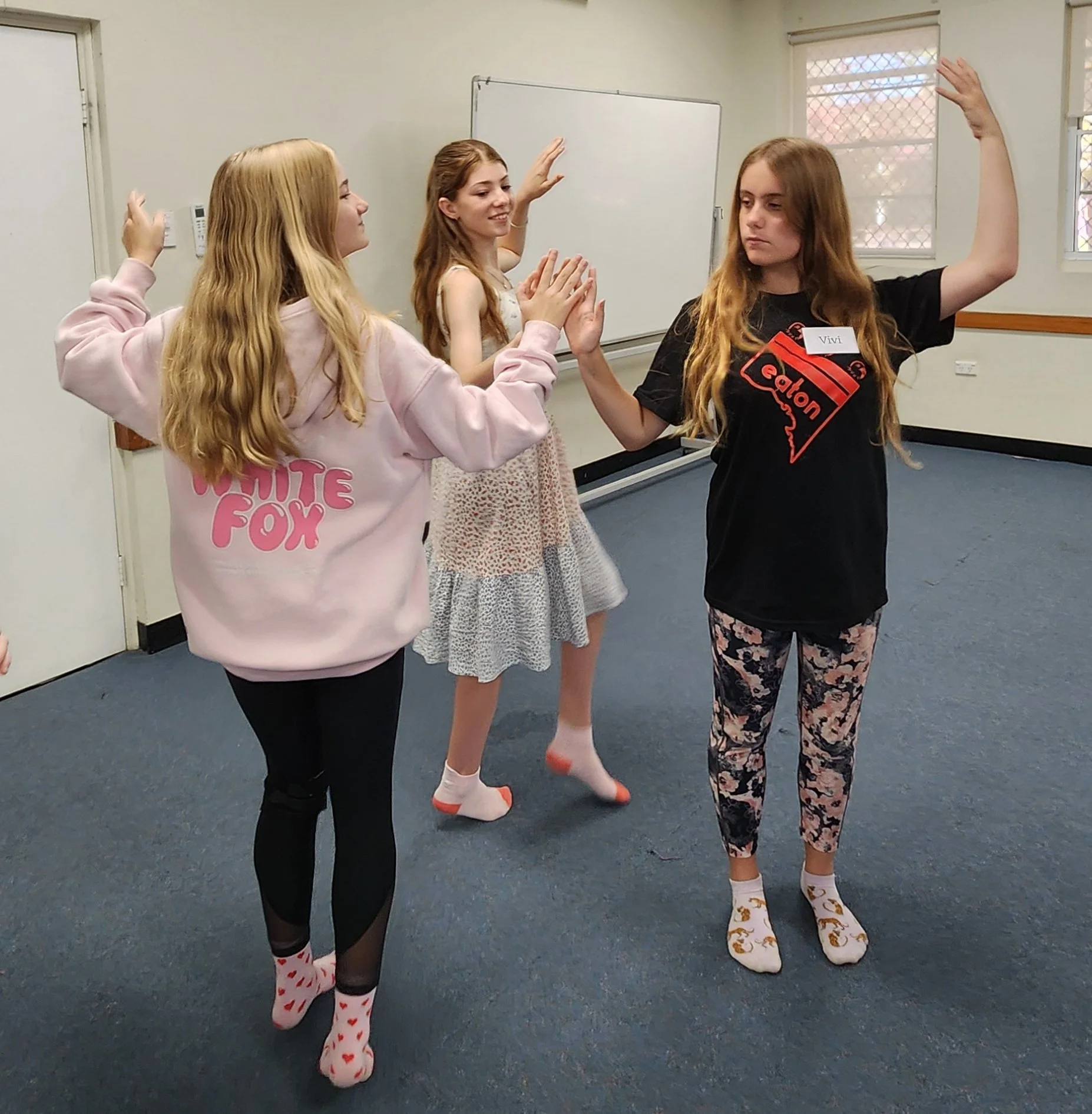 Four girls in a room with blue carpet, engaging in a playful activity of high-fiving each other. They are dressed casually, and one girl is wearing pajamas with heart patterns. Three of the girls have long, light-brown or red hair. The room has windows with mesh screens, a whiteboard, and beige walls.