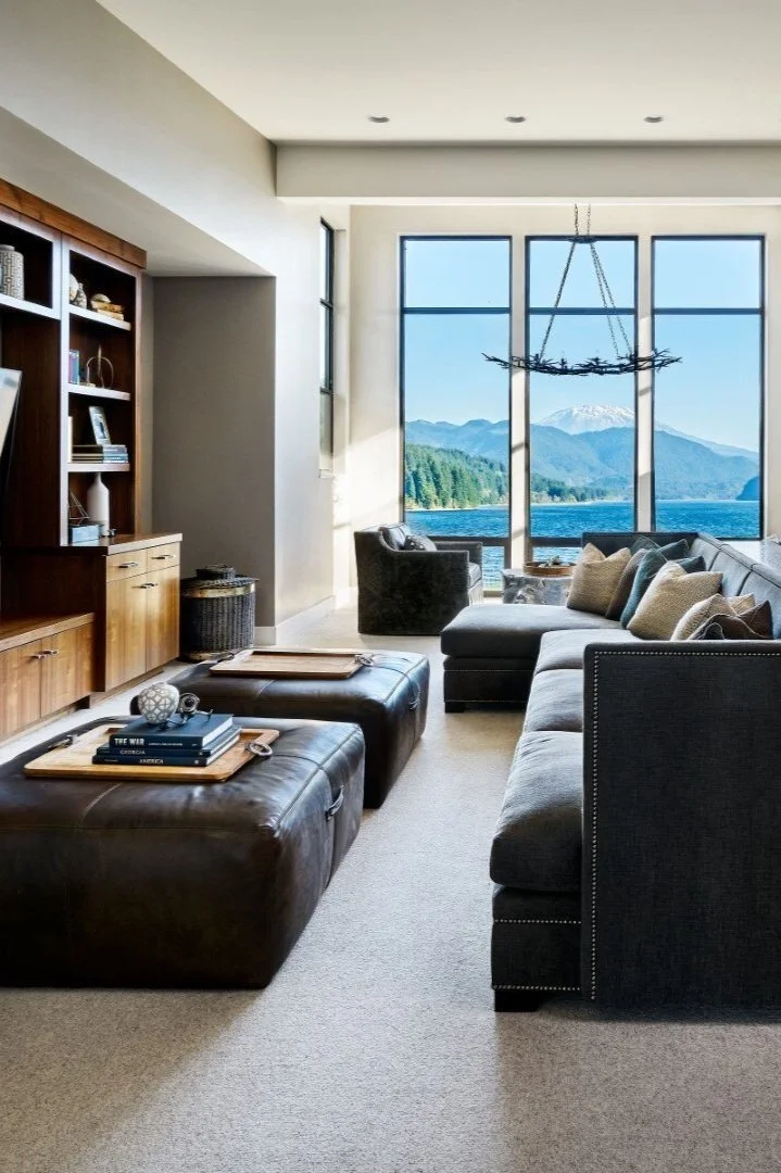 Modern living room with large windows overlooking a scenic lake and mountain landscape, featuring a dark gray sectional sofa, leather ottomans, wooden shelves with decorative items, and a rustic chandelier.