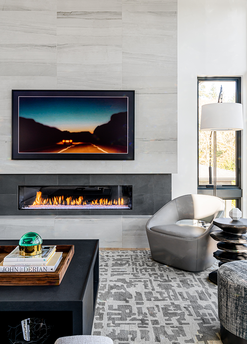 Modern living room with a wall-mounted flat-screen TV above a linear gas fireplace, a white modern armchair near a window with a floor lamp, and a black coffee table with books and decorative items, all in a bright space with neutral tones and a patterned area rug.