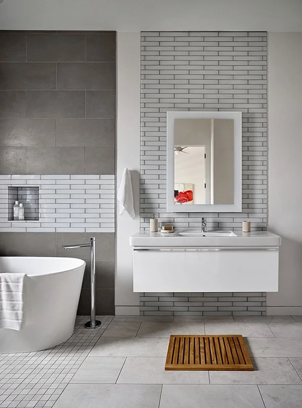 Modern bathroom with a white freestanding bathtub, a wall-mounted white vanity with a mirror, and a gray and white tiled wall with a built-in shelf.