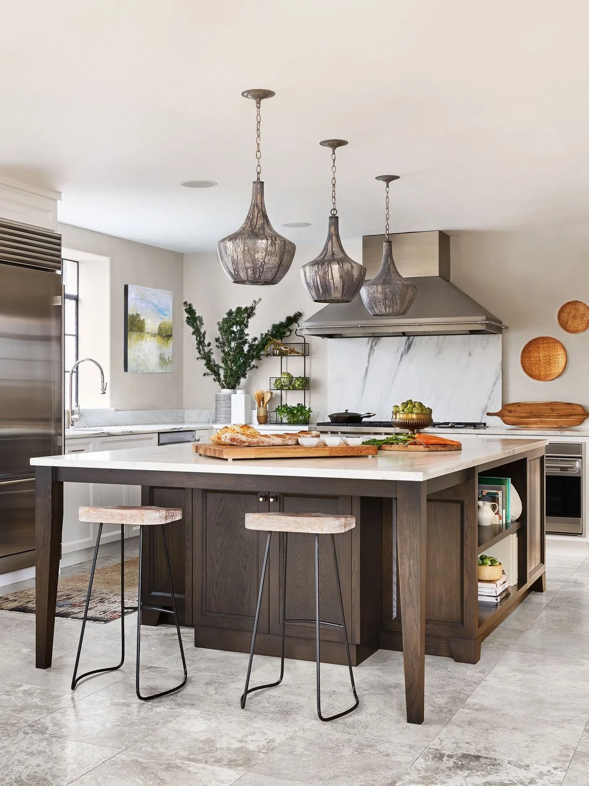 A modern kitchen with a large island, pendant lights, stainless steel appliances, open shelving with copper and glassware, and decorative baskets on the wall.