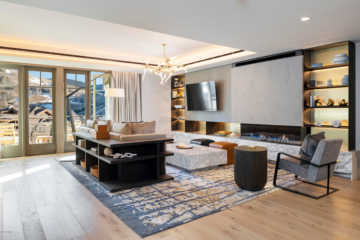 Modern living room with large windows, mountain view, abstract chandelier, TV, fireplace, shelves with decor, patterned rug, armchair, and seating area.