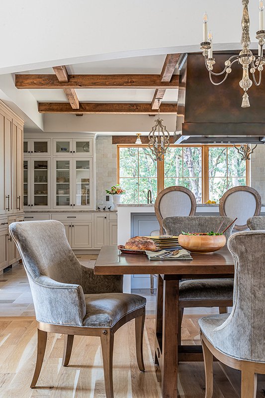 Dining area with a wooden table, upholstered chairs, a chandelier, and large windows showing greenery outside in a bright, modern kitchen.