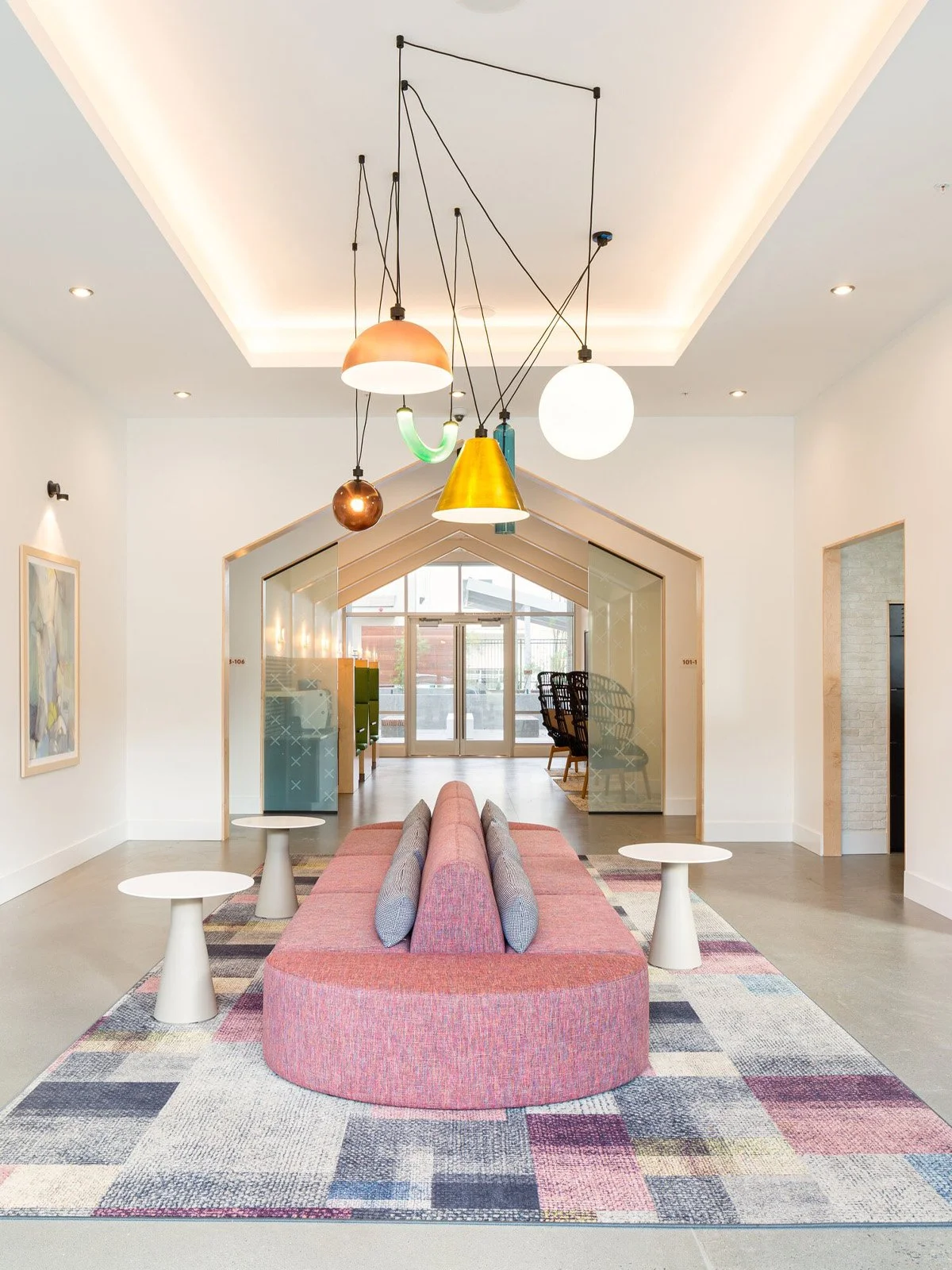 Modern lobby area with a pink upholstered curved bench, white side tables, a colorful geometric patterned rug, and hanging pendant lights in various shapes and colors ceiling.