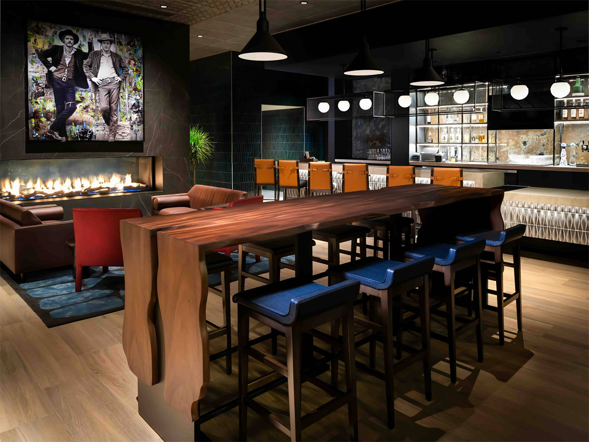 Modern bar and lounge area with a long wooden table, high chairs with blue upholstered seats, a fireplace, and a bar with orange chairs, illuminated by hanging pendant lights.