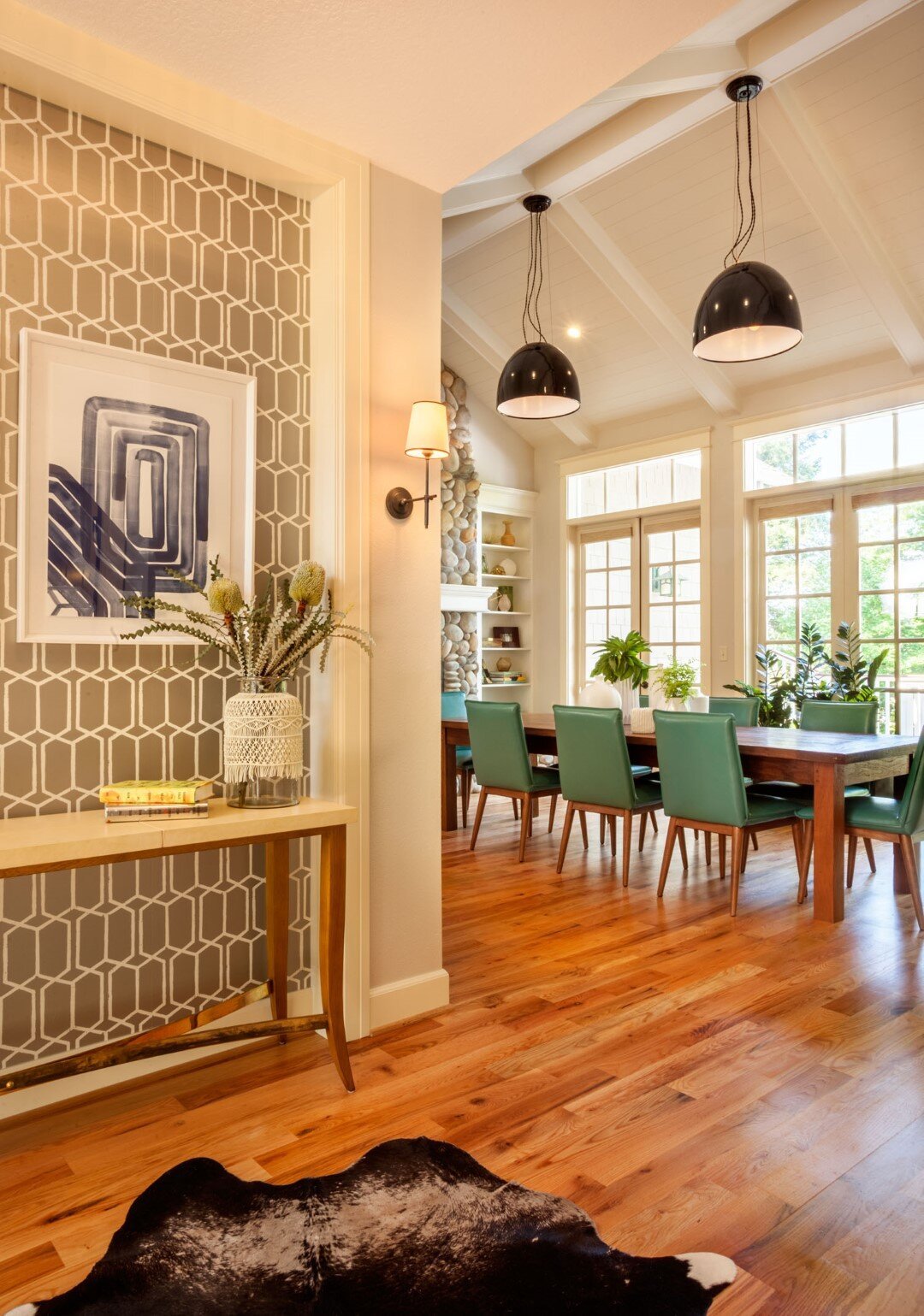 Contemporary dining room with a wooden floor, turquoise chairs, a large wooden table, and pendant lights. Decorative wall with a geometric pattern, a table with a plant and books, and animal print rug on the floor.