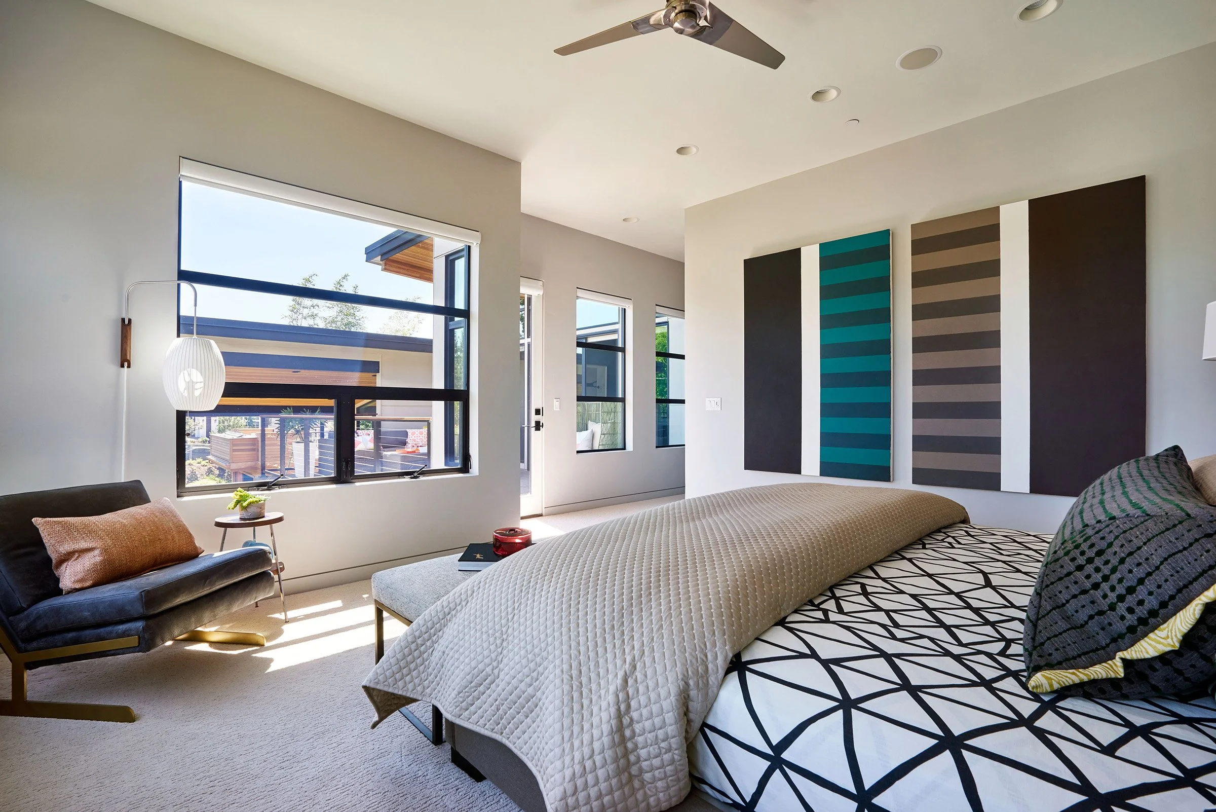 A bedroom with large windows, decorative bedding and a reading chair.