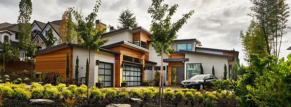 Modern residential homes with large windows, surrounded by lush landscaping and trees, with a black car parked outside.