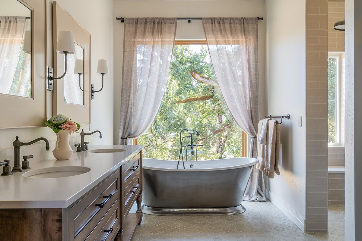 Bathroom with a freestanding bathtub by a large window with beige curtains, a double vanity with two sinks, a vase with flowers, and wall-mounted lights.