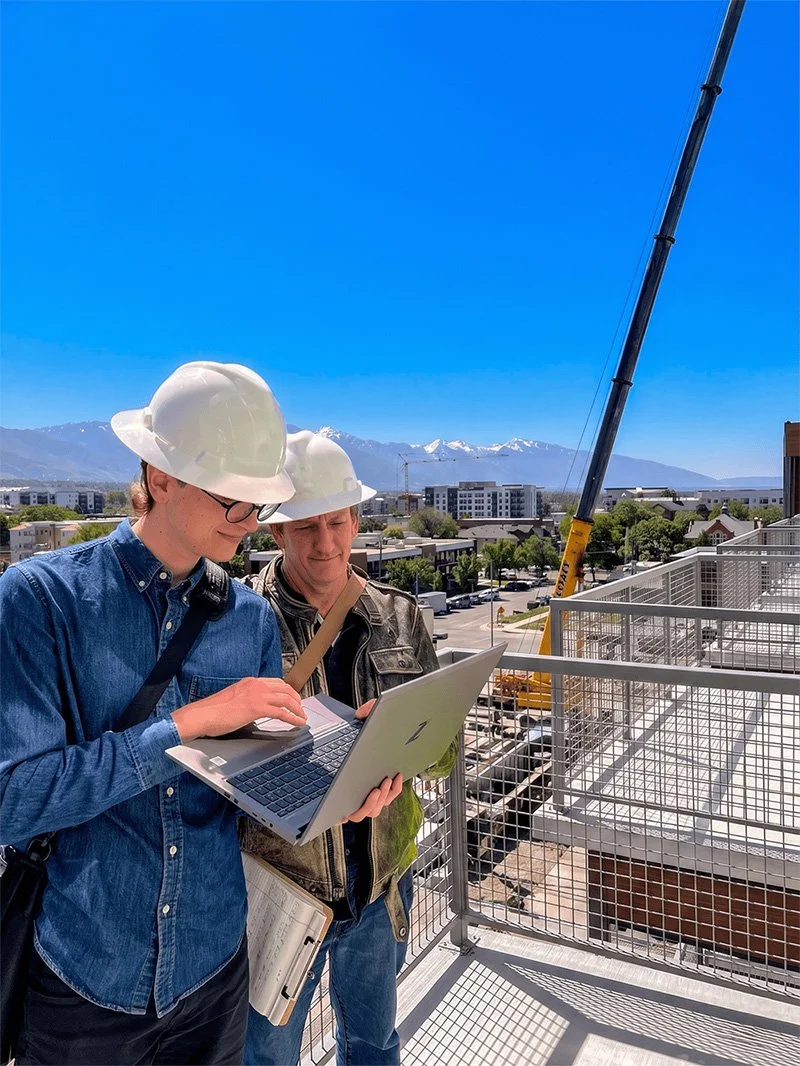Two construction workers wearing white safety helmets standing on a balcony of a building, looking at a laptop, with a crane in the background and snow-capped mountains under a clear blue sky.