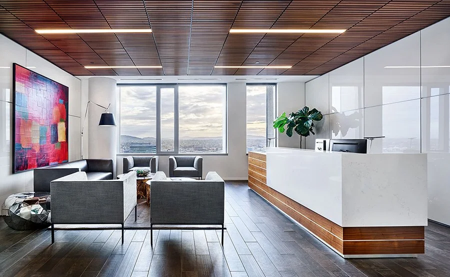 Modern office lounge with gray chairs, a black couch, abstract art, large windows showing cityscape, and a reception desk with a potted plant.