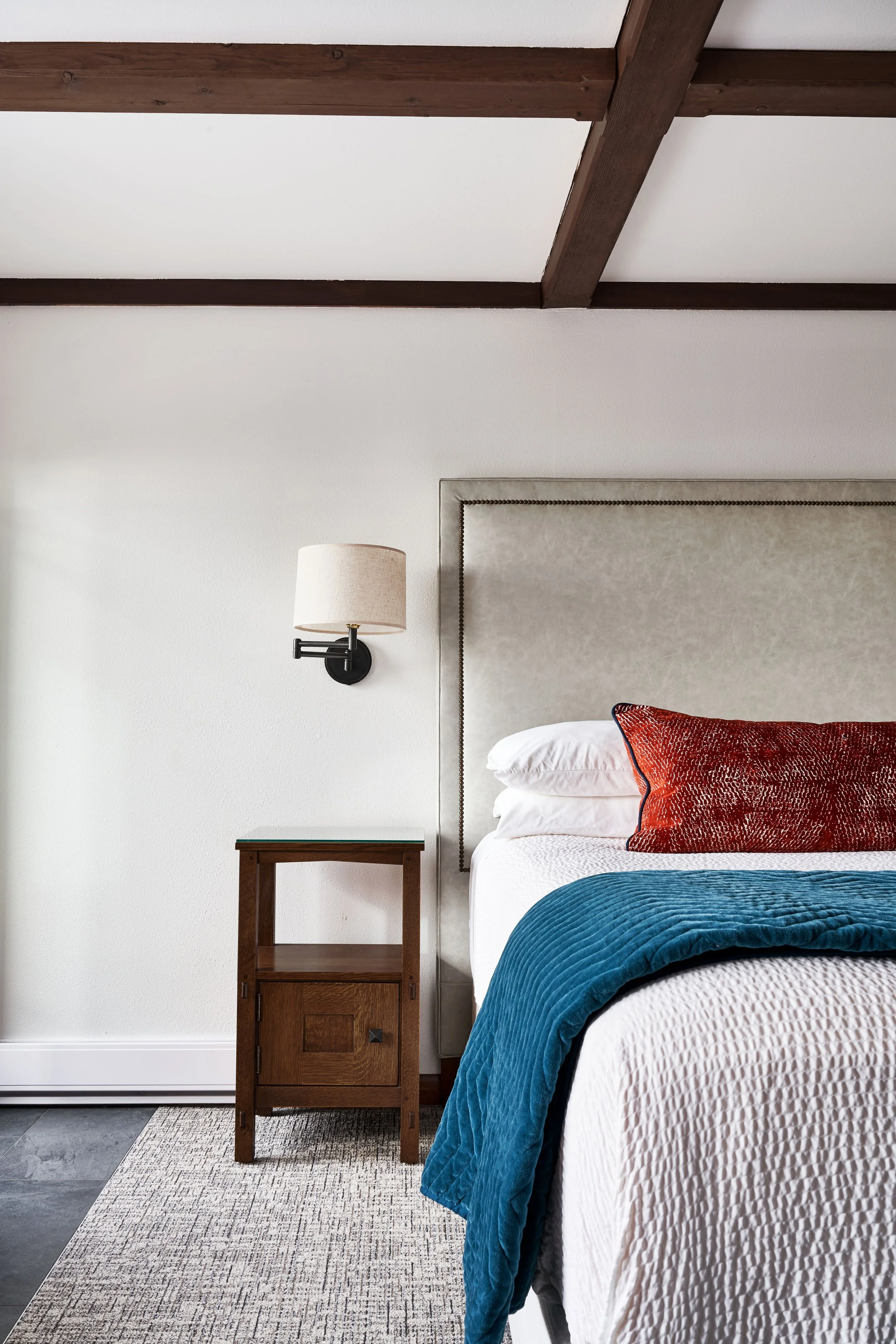 Bedroom interior with a bed featuring a white mattress and blue quilt, red decorative pillow, wooden nightstand, wall-mounted lamp, and exposed ceiling beams.