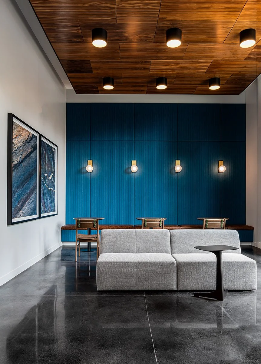 Modern interior lounge with a gray sofa, small black side table, blue accent wall with four hanging lights, wooden ceiling with recessed lighting, and abstract art on the white wall.