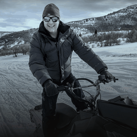 Person in winter clothing riding a snowmobile on a snowy landscape with hills and trees in the background.