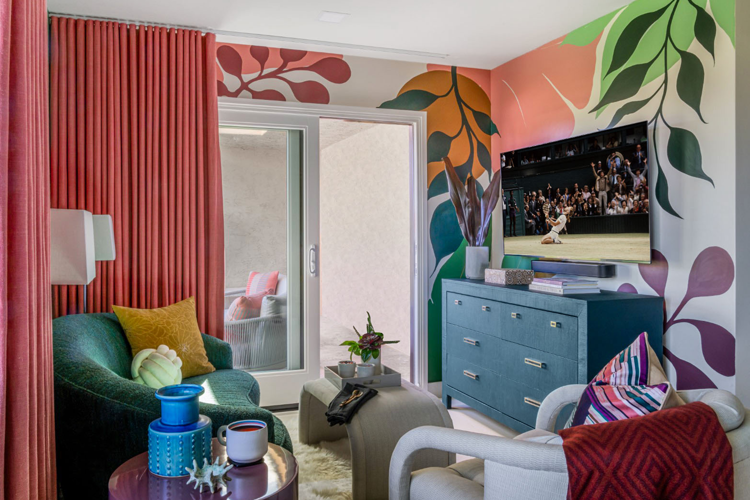Palm Desert condo TV niche after remodel, bold botanical mural with curated vintage seating and wall-mounted TV, by Studio Garrison.