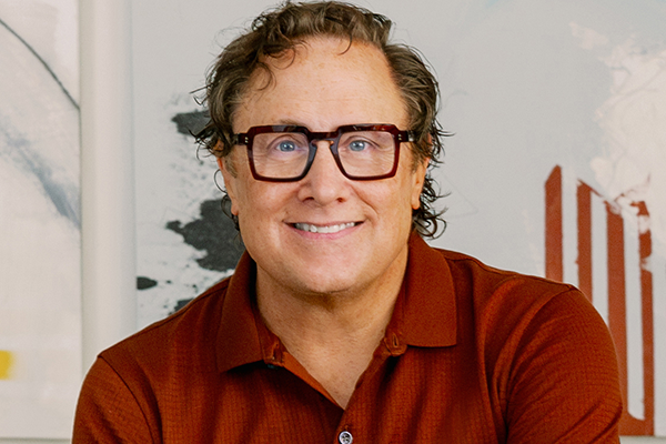 A man with curly hair and glasses smiling, wearing a reddish-brown polo shirt