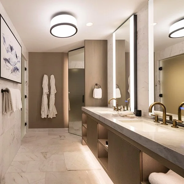 Modern bathroom with double vanity, marble surfaces, large mirrors, gold faucets, walk-in shower, robes, and abstract wall art.