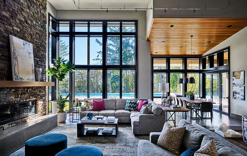 Modern living room with large floor-to-ceiling windows, a brick fireplace, a light-colored sectional sofa, colorful pillows, a dark wooden coffee table, and a blue ottoman. Adjacent dining area with a wooden ceiling, black dining table, and pendant lighting, overlooking an outdoor pool.