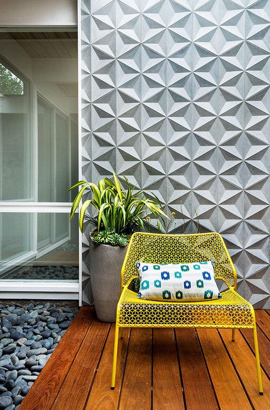 A yellow outdoor chair with a patterned cushion, next to a large potted plant, on a wooden deck, with a textured geometric patio wall and a glass window in the background.