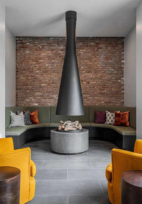 A modern lounge area with a circular concrete fire pit in the center, surrounded by a curved green and black sectional sofa adorned with colorful pillows, a brick accent wall in the background, and two yellow armchairs in the foreground.