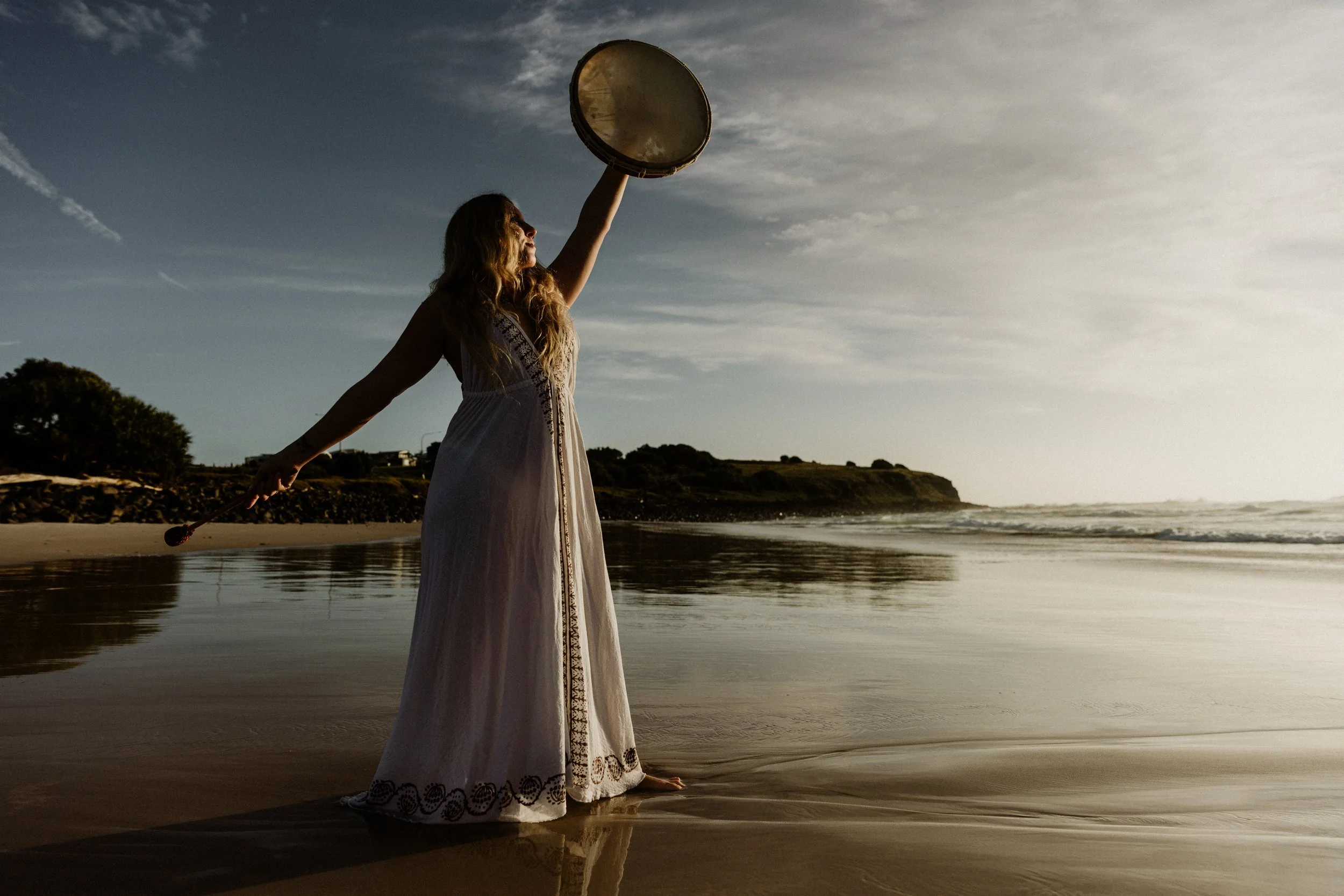 A woman wearing a long white dress standing barefoot on the beach, holding a large drum above her head while facing the ocean during sunset or sunrise.