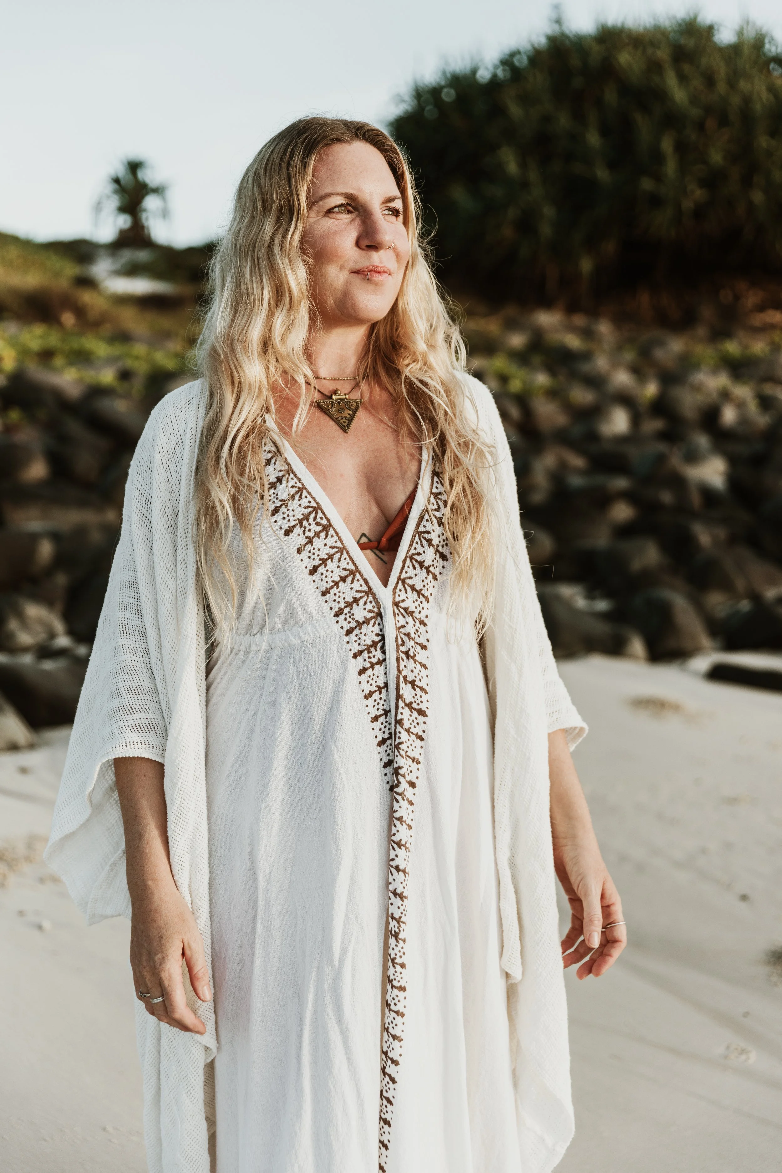 A woman with long, wavy blond hair standing on a beach, wearing a white dress and a woven necklace with a triangle pendant, gazing into the distance with dry rocky terrain and some greenery in the background.