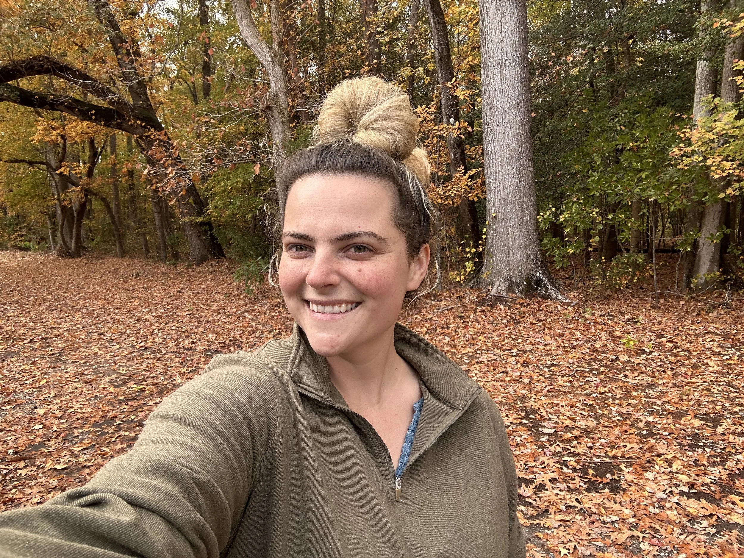 A woman taking a selfie outdoors in an autumn forest with fallen leaves on the ground and trees with colorful fall foliage.