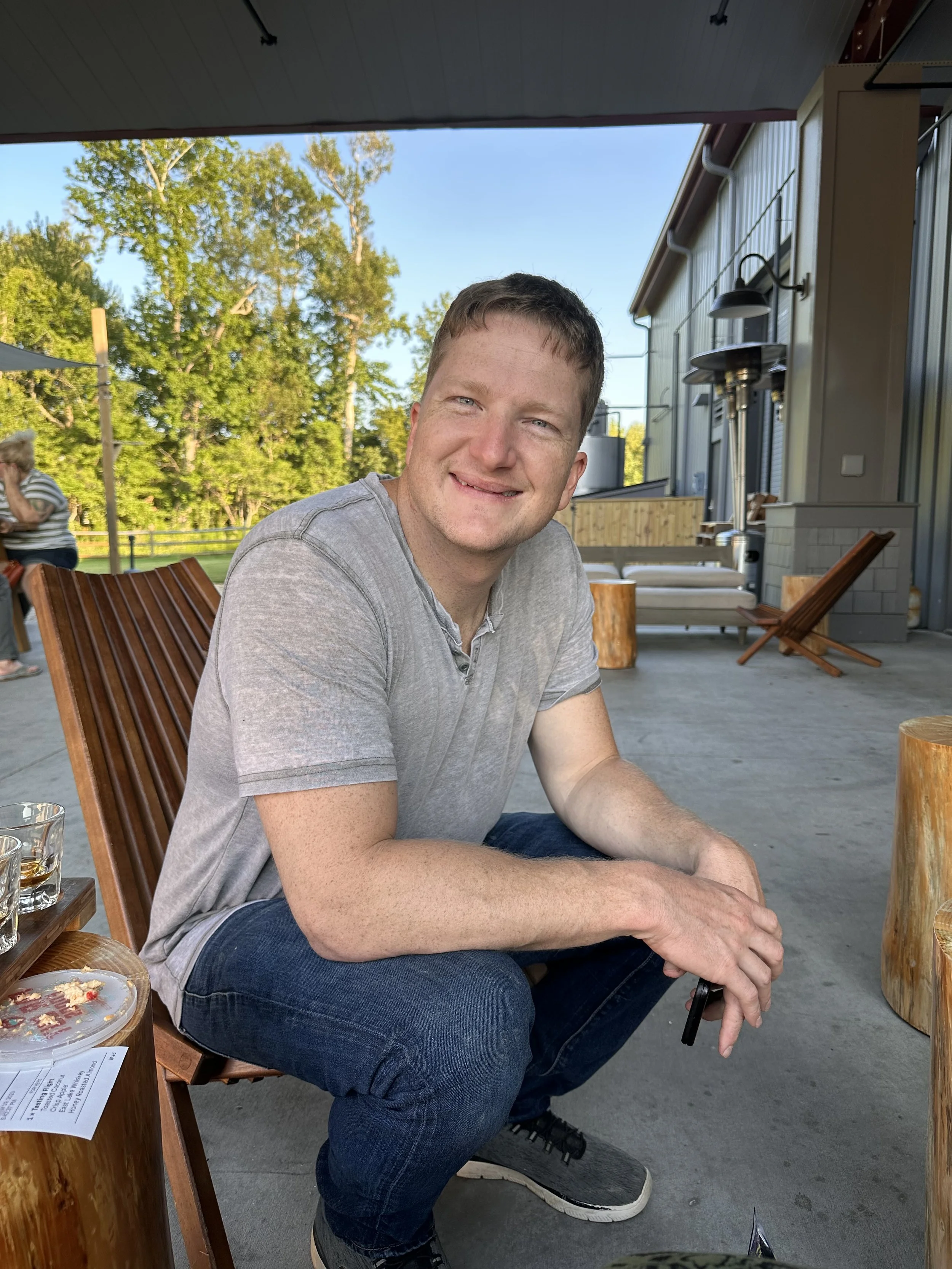 A young man with short brown hair and blue eyes smiling at the camera, sitting outdoors on a wooden chair with a relaxed posture, with trees and a building in the background.