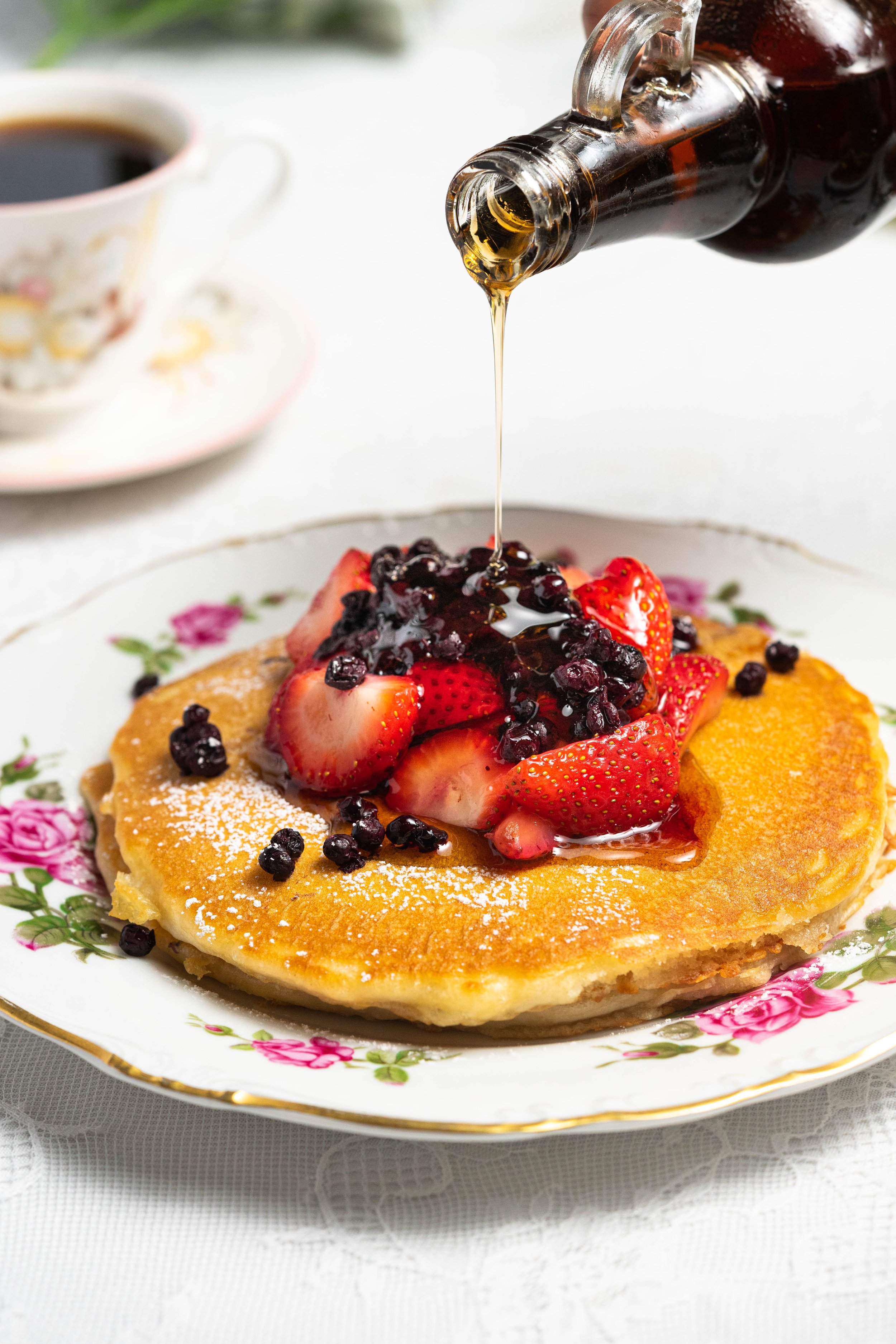 Pancakes topped with strawberries and berries, drizzled with syrup, on a floral plate, with a cup of coffee in the background.