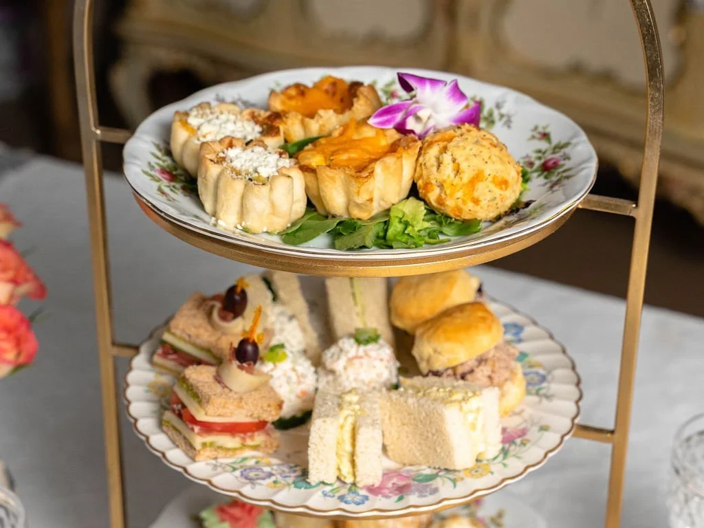 Three-tiered stand with assorted pastries, quiches, sandwiches, and scones on decorative plates for afternoon tea.